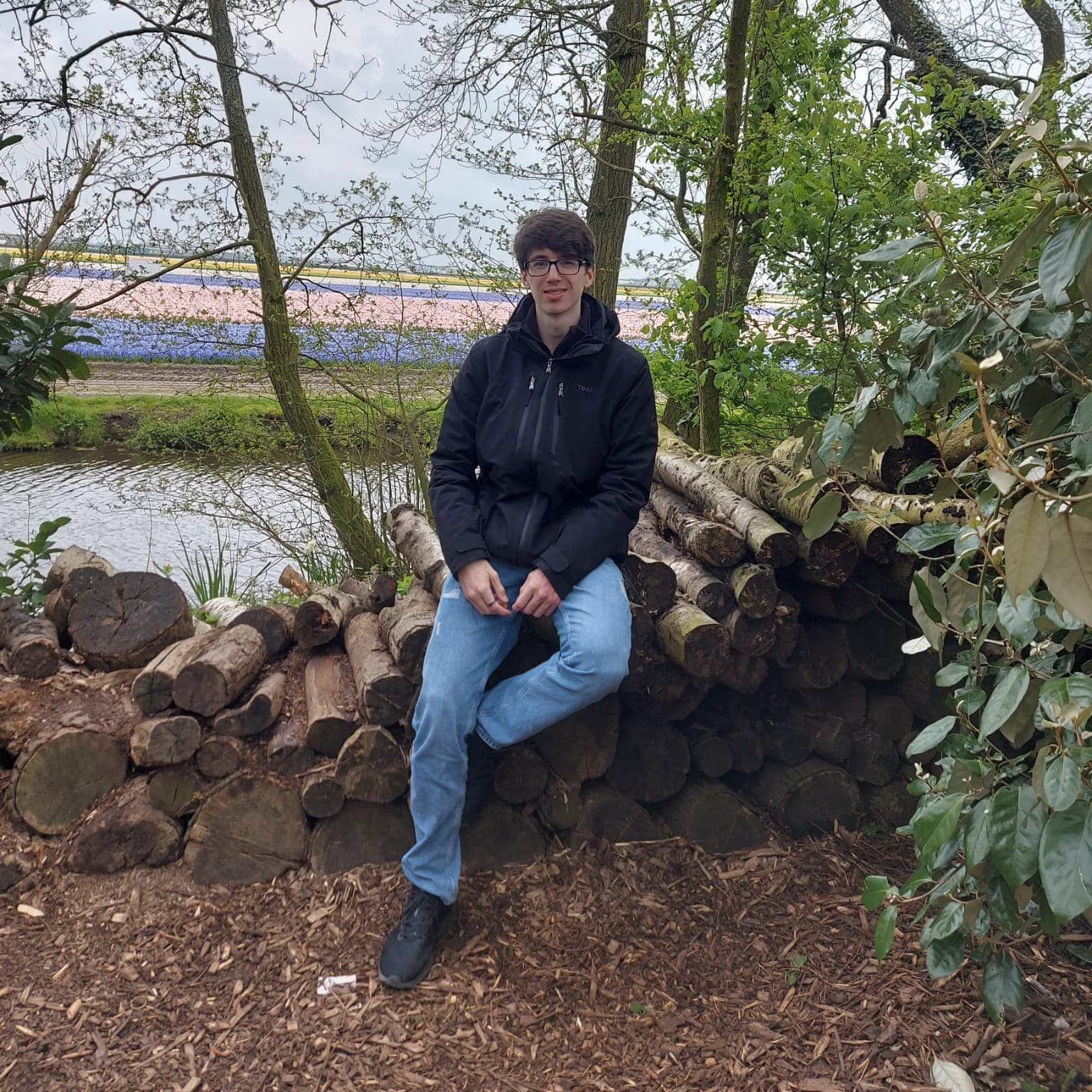 A photo of young, brown-haired man (me), sitting on a pile of logs in front of a field of tulips.